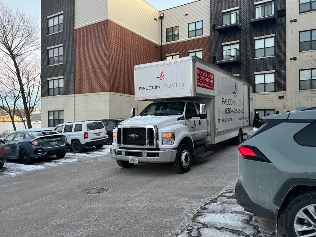A white box truck parked in front of a modern multi-story apartment building with a partially snow-covered parking lot.
