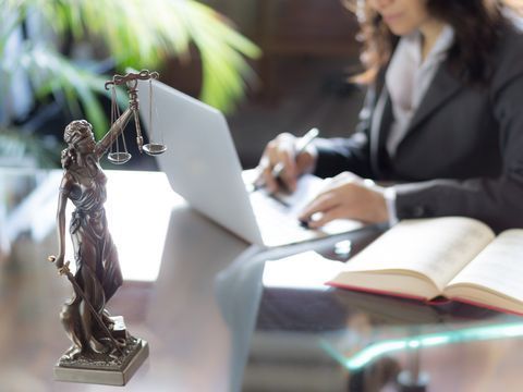 A woman is sitting at a desk with a laptop and a statue of justice.