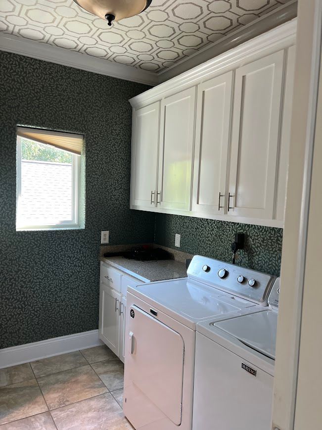 Laundry room with white cabinets, washer, and dryer. Green patterned walls, patterned ceiling, and window.