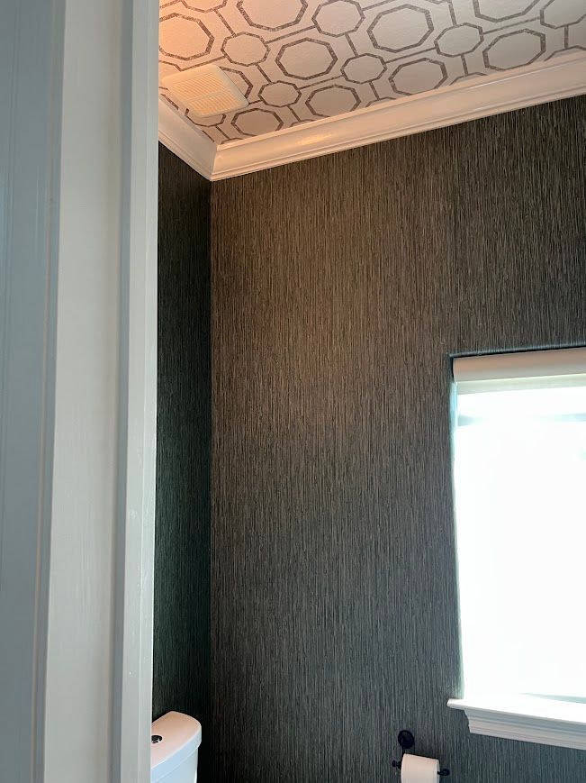 Dark, textured wallpaper in a bathroom, patterned ceiling, white trim, and a window with a closed blind.