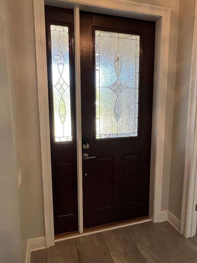 Brown front door with decorative glass panel and sidelight, white trim, wood-look floor.