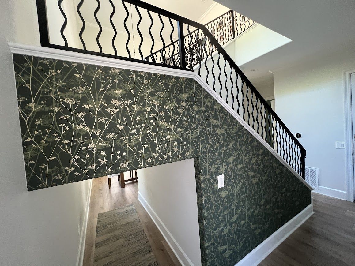 Staircase with dark floral wallpaper. Black iron railing with white trim. Wooden floor and a small area rug.