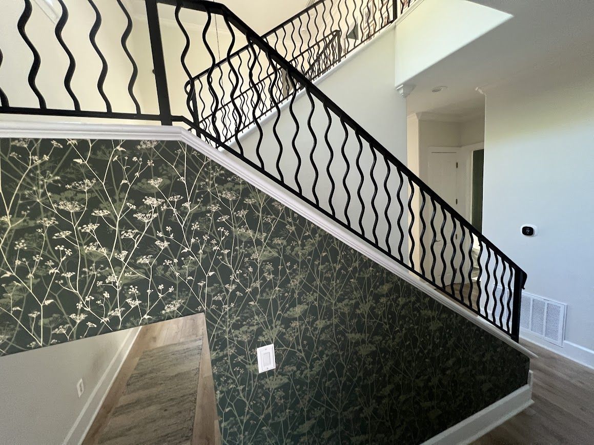 Staircase with black wavy metal railing, green and white patterned wallpaper, and wooden floors.