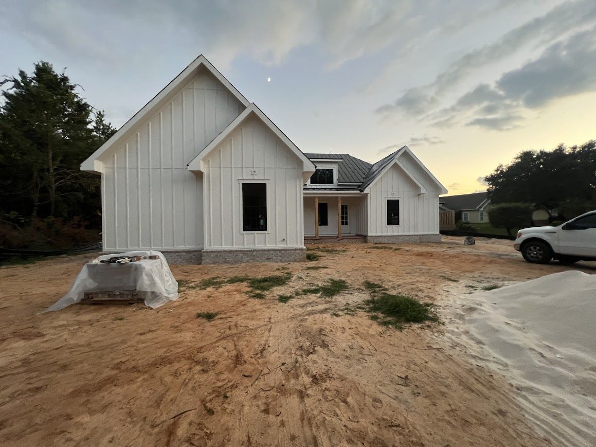 White house under construction, with dark window frames, set on a sandy lot under a cloudy sky.