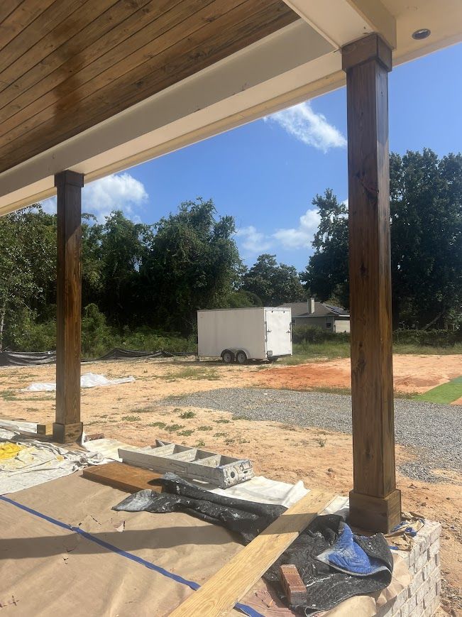 Wooden porch posts frame a view of a dirt lot with construction materials, trees, and a white shed under a blue sky.