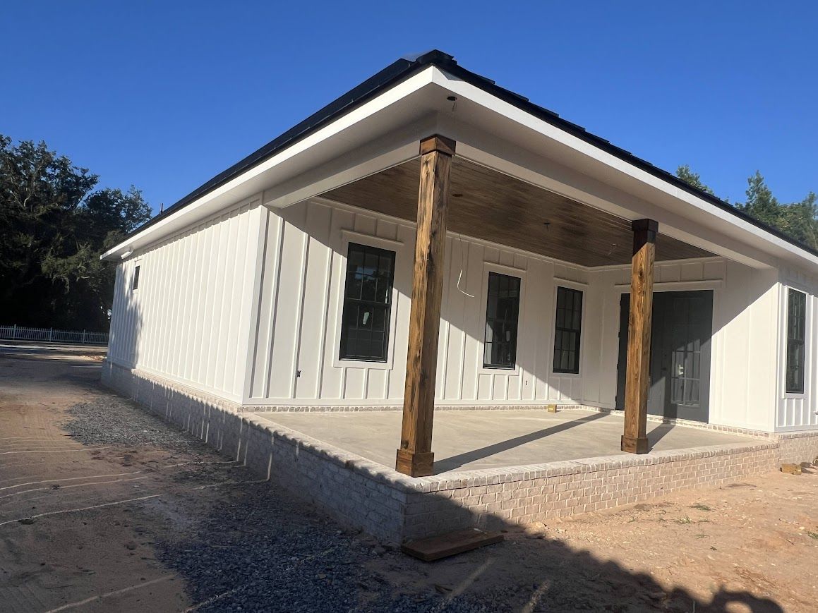 White-sided house with a porch supported by brown wooden posts, under a black roof on a sunny day.