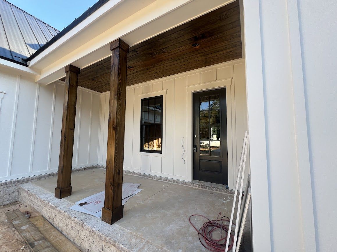 Porch with dark wood columns, white siding, black door, and unfinished concrete floor.