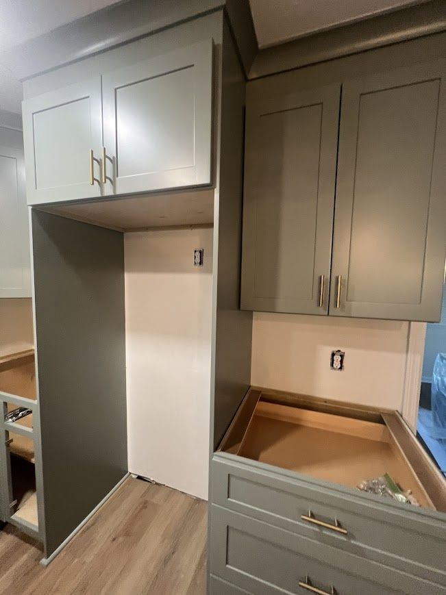 Kitchen cabinets in varying stages of completion; gray-green color. A refrigerator space is framed by the cabinet.