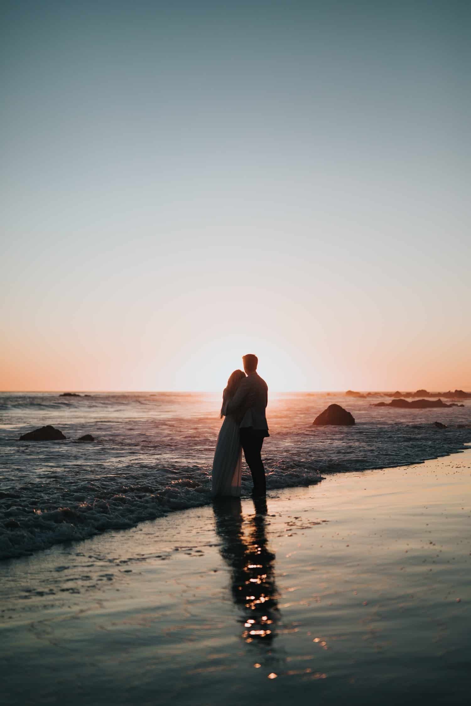 A man and woman are hugging on the beach at sunset.
