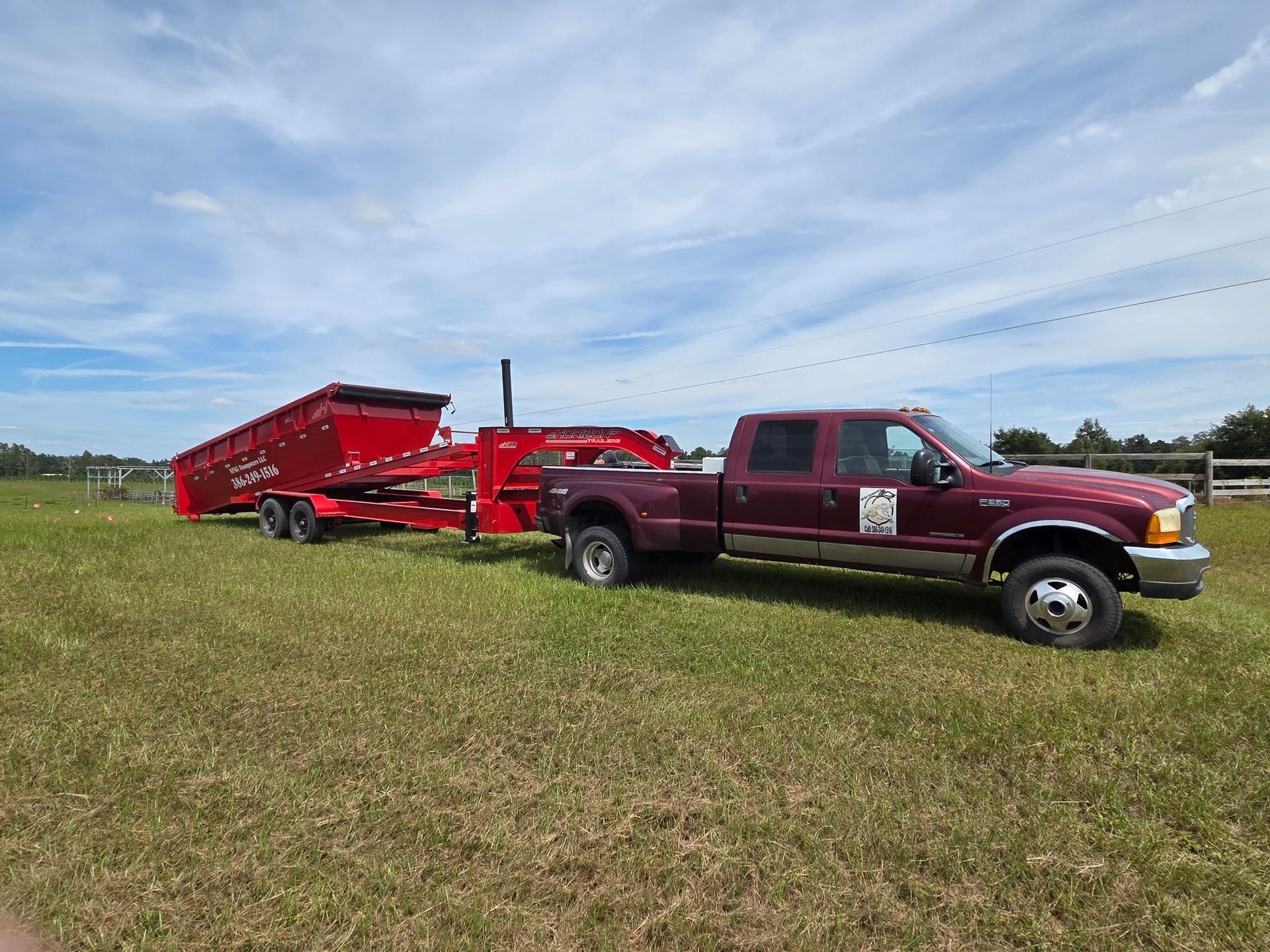 Red truck towing a red dump trailer on grass field under a partly cloudy sky.