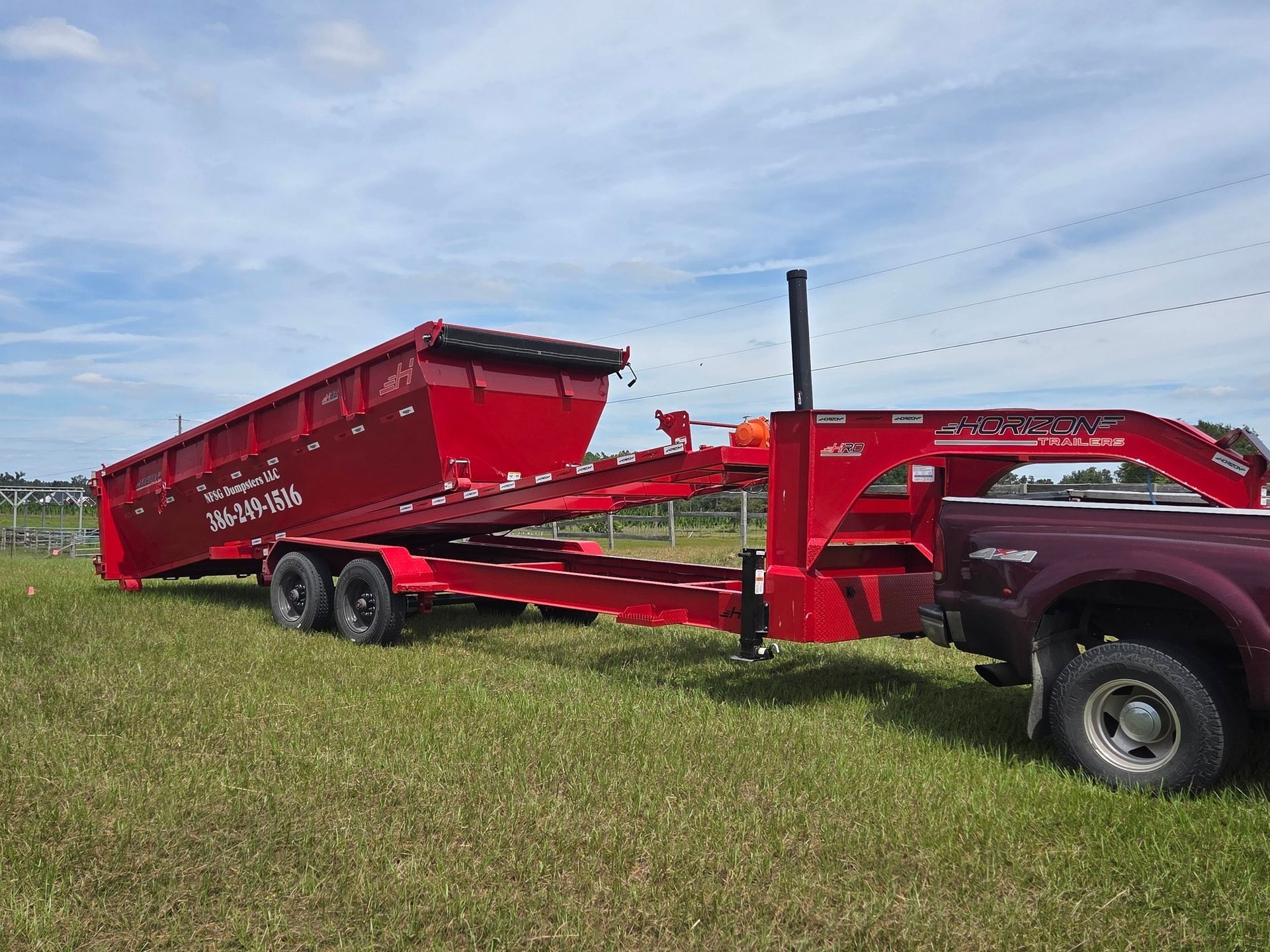 Red roll-off dumpster trailer hooked to a truck on a grassy field; trailer tilted upwards.