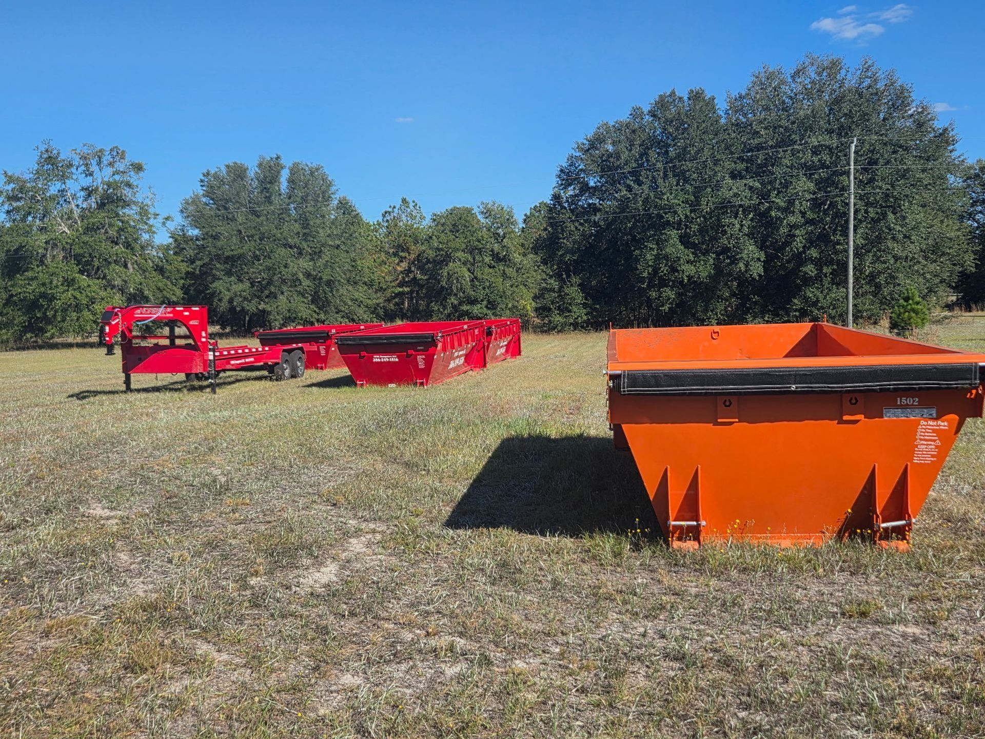 Orange dumpsters and a trailer on a grassy field with trees under a blue sky.