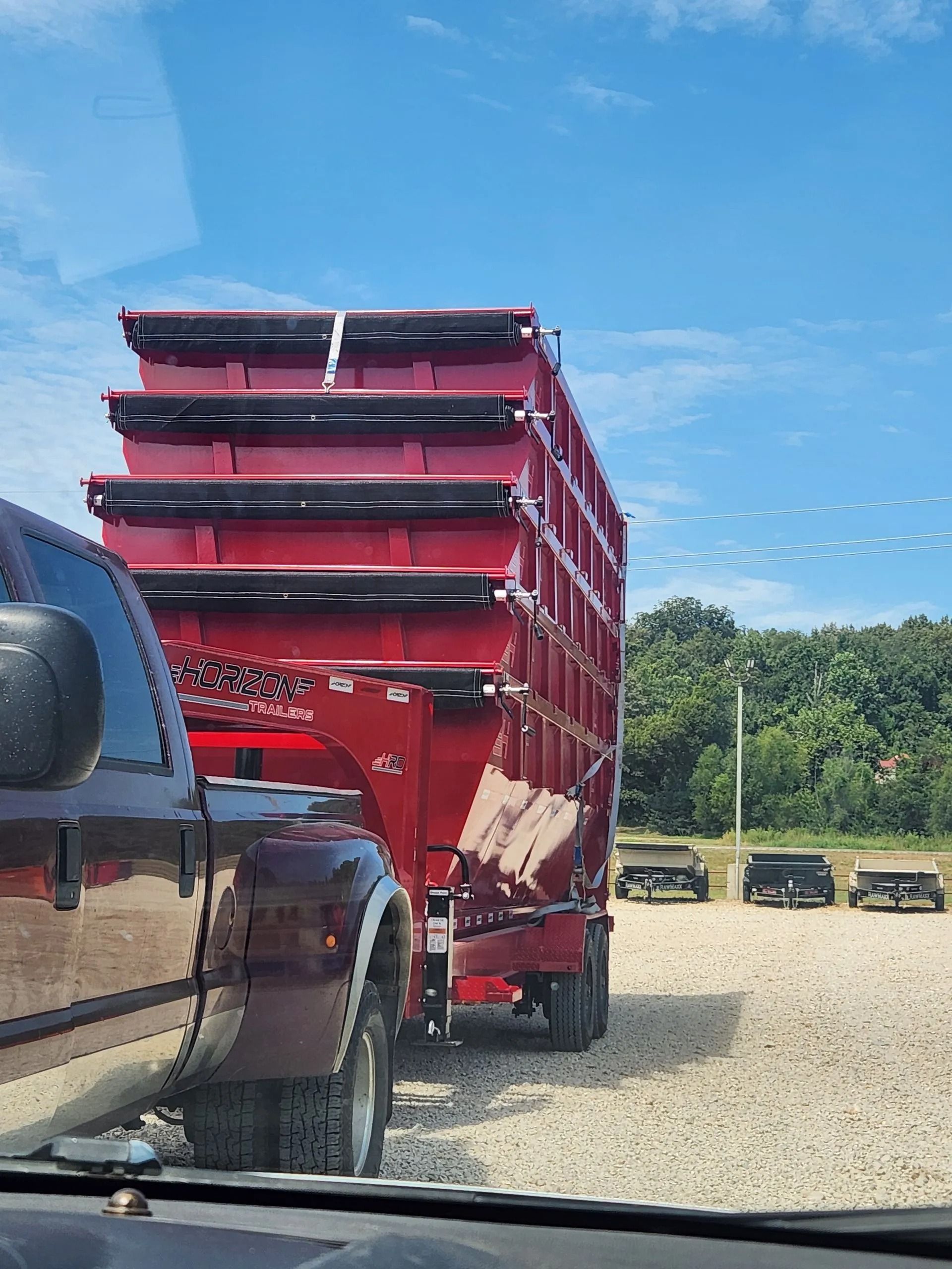 Red grain trailer hitched to a truck, parked on gravel in front of trees under a blue sky.