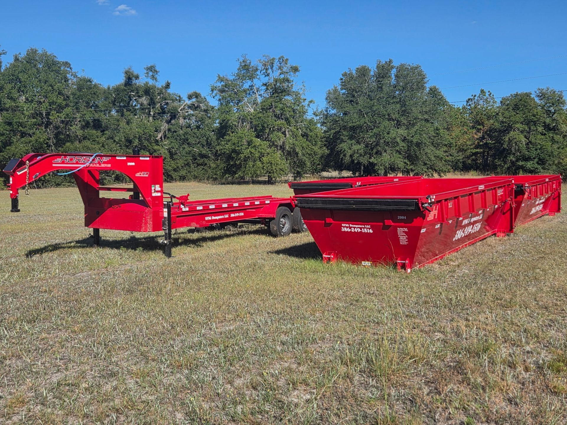 Red dump trailer in a grassy field with trees under a blue sky.