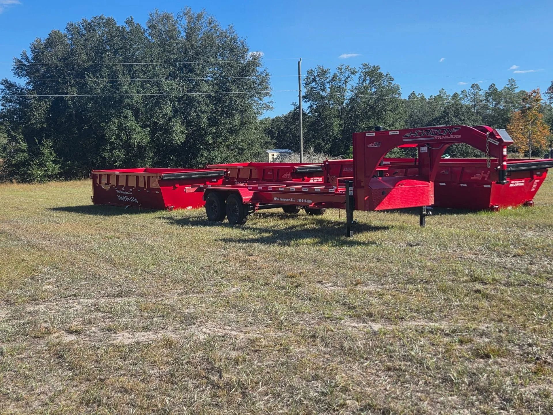Red roll-off trailer and two containers in a grassy field under a blue sky.
