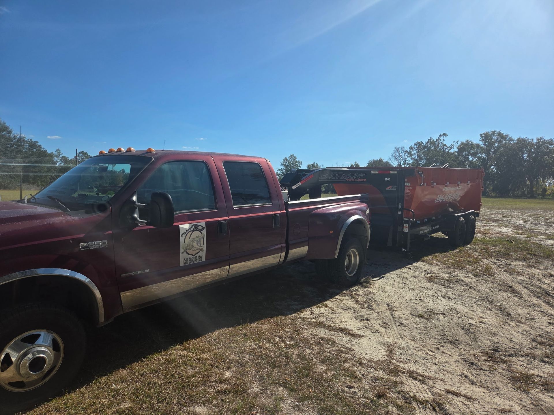Burgundy Ford truck with a trailer parked on a sandy area under a bright blue sky.