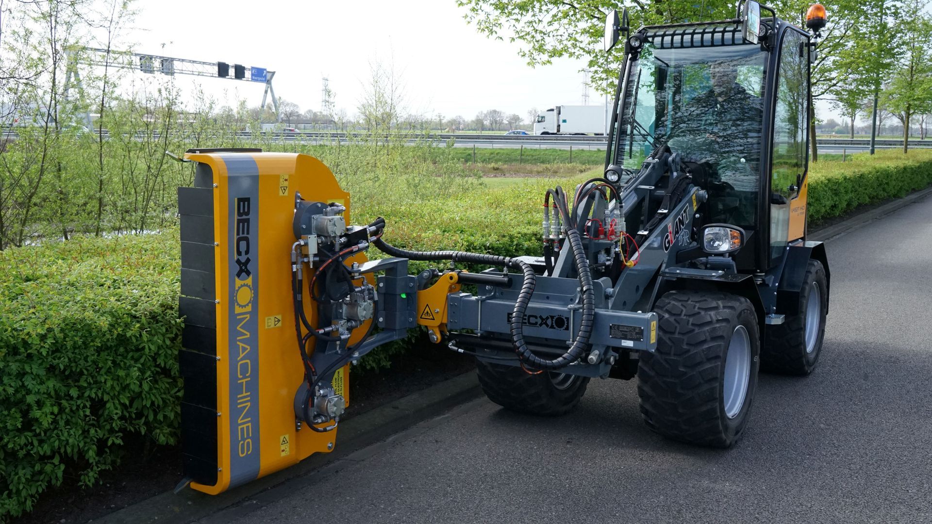 Ein gelb-schwarzer Traktor steht am Straßenrand neben einer Hecke.