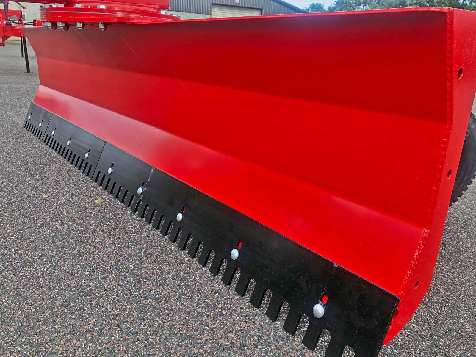 Auf einer Schotterstraße steht ein roter Schneepflug mit schwarzen Zähnen.