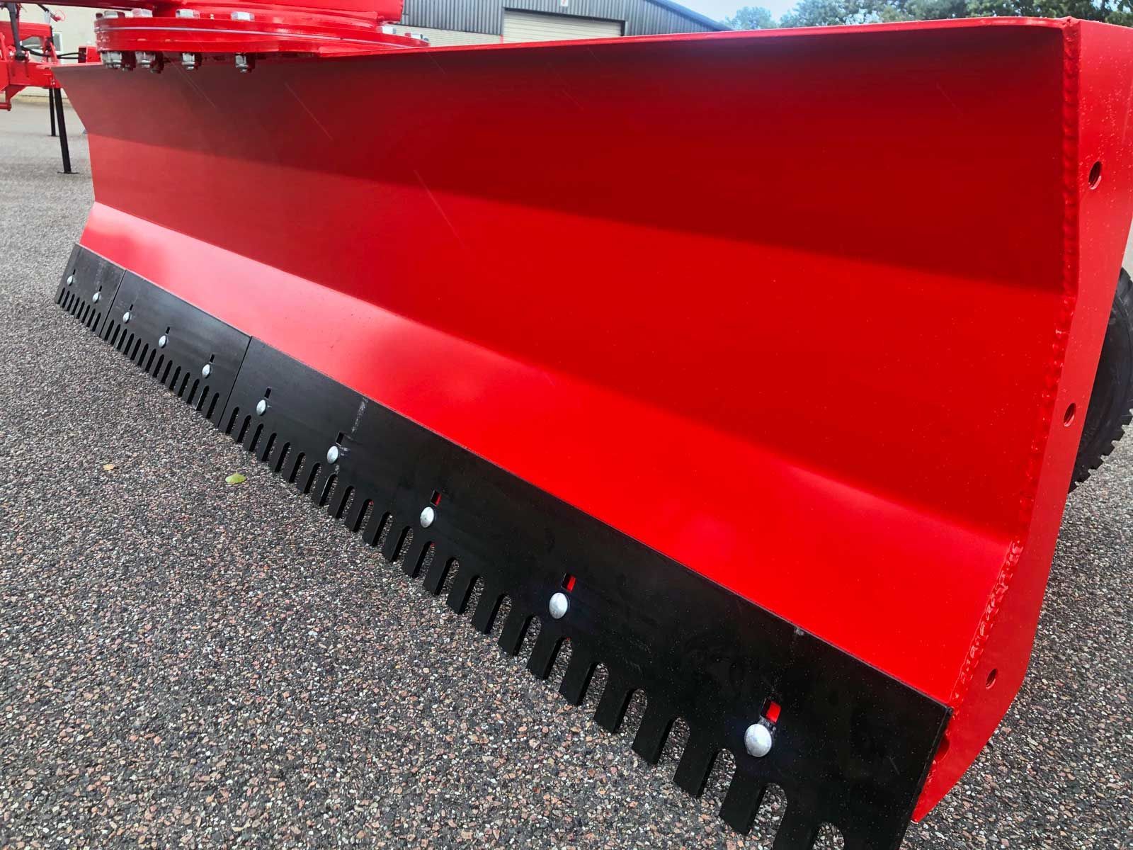 Auf einer Schotterstraße steht ein roter Schneepflug mit schwarzen Zähnen.