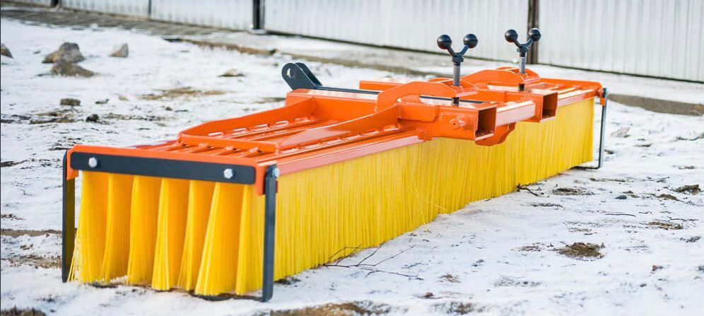 Orangefarbener und gelber Bürstenaufsatz für einen Traktor, der auf schneebedecktem Boden steht.