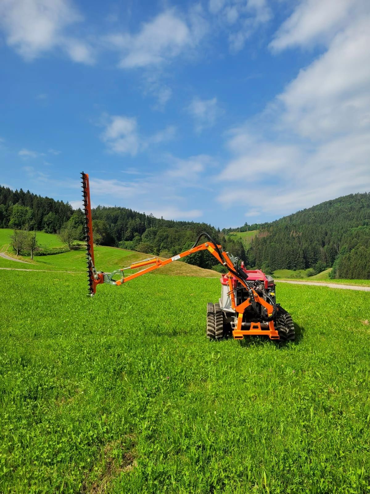 Ein orangefarbener Traktor mit ausgefahrenem Mäharm mäht Gras auf einer grünen Wiese unter blauem Himmel.