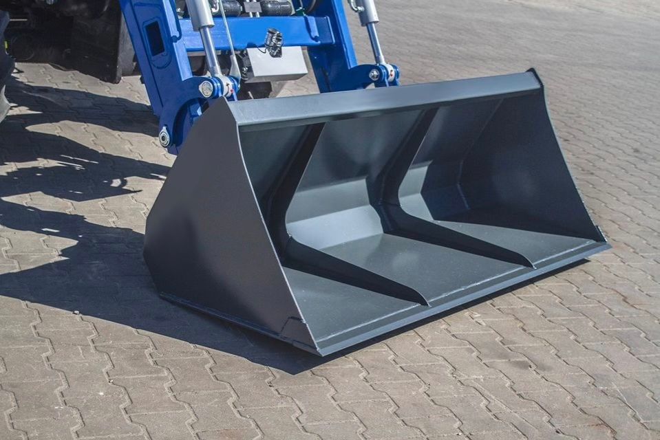 Auf einem Ziegelsteinpflaster steht neben einem blauen Traktor ein schwarzer Eimer.