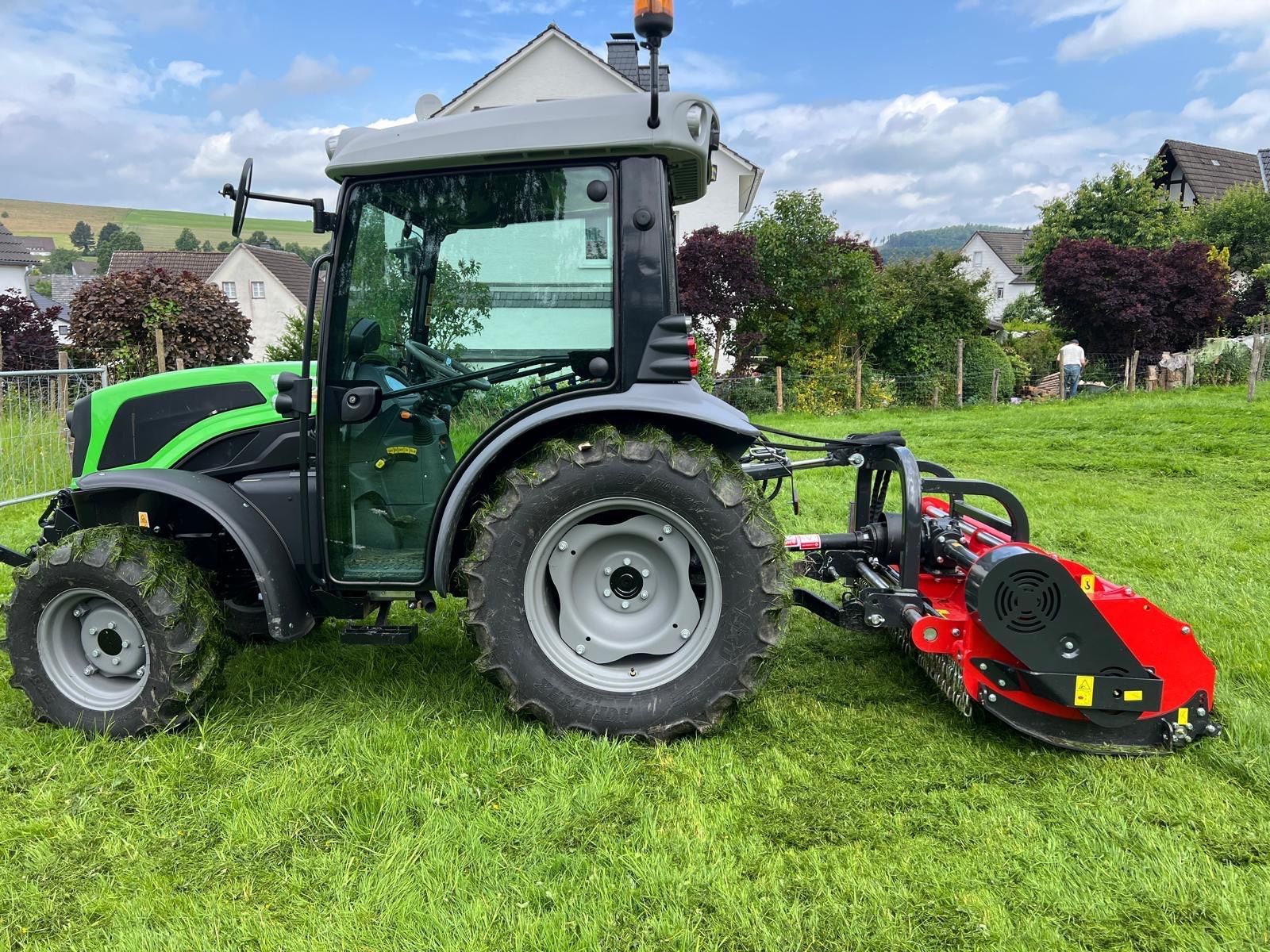 Ein grüner Traktor mit einem daran montierten roten Mäher steht auf einer Wiese.
