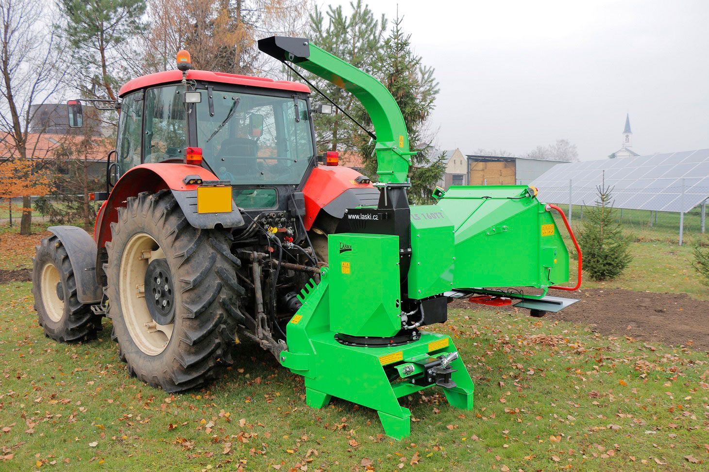Ein roter Traktor mit einem daran befestigten grünen Häcksler steht auf einer Wiese.