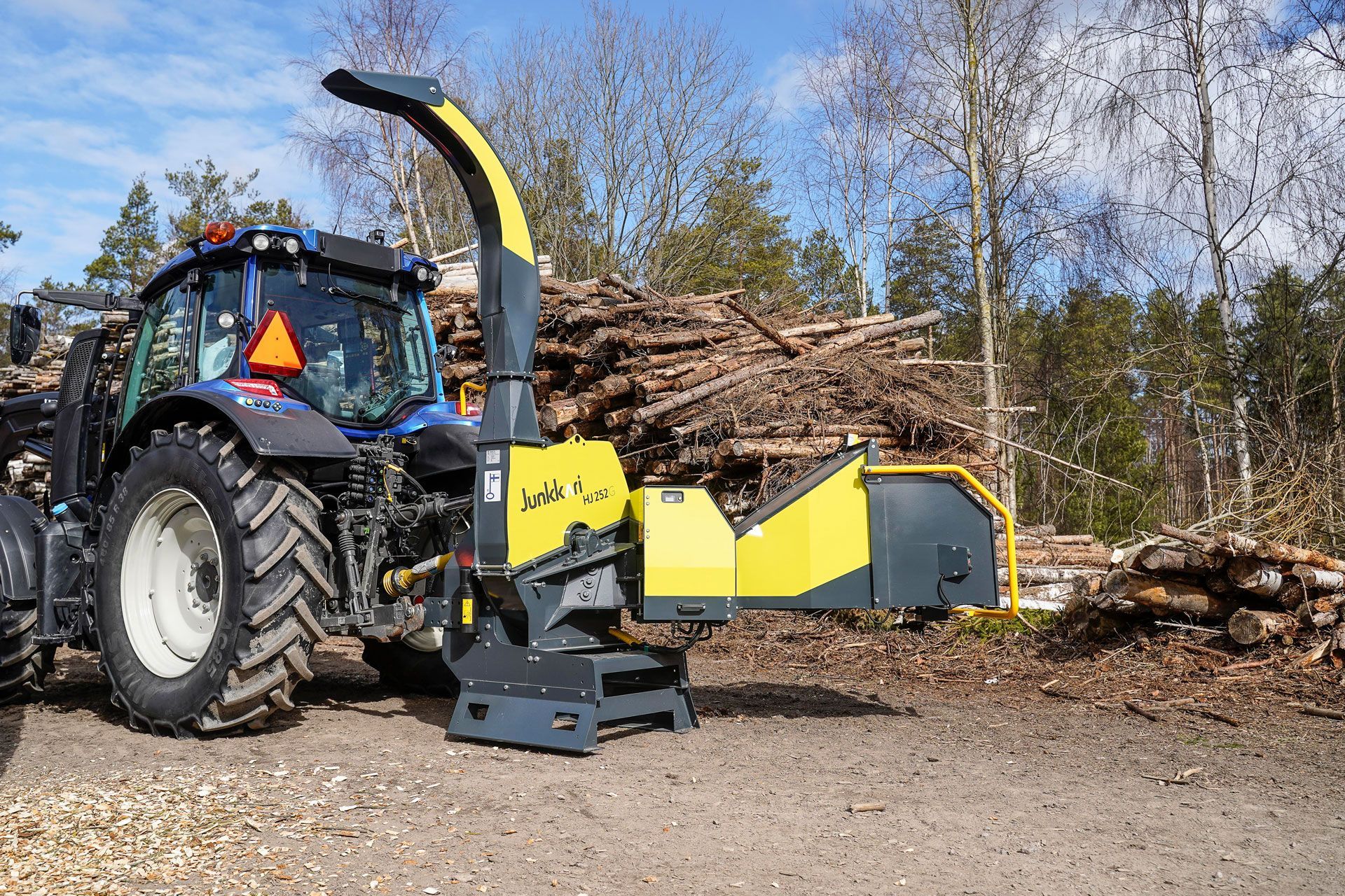 Ein gelb-schwarzer Traktor häckselt Holz vor einem Stapel Holzstämme.