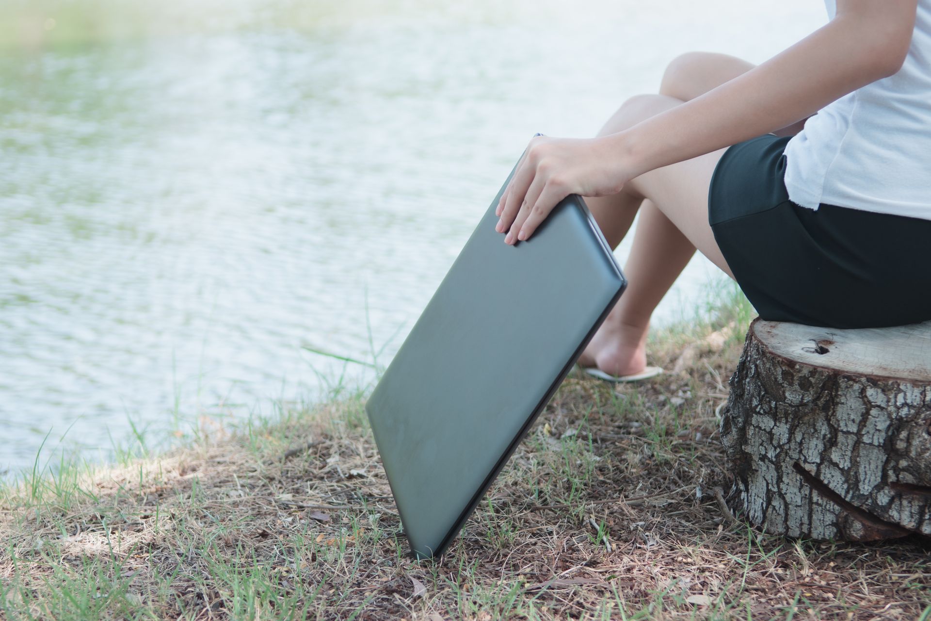 Woman holding laptop, sitting on tree stump by a body of water.