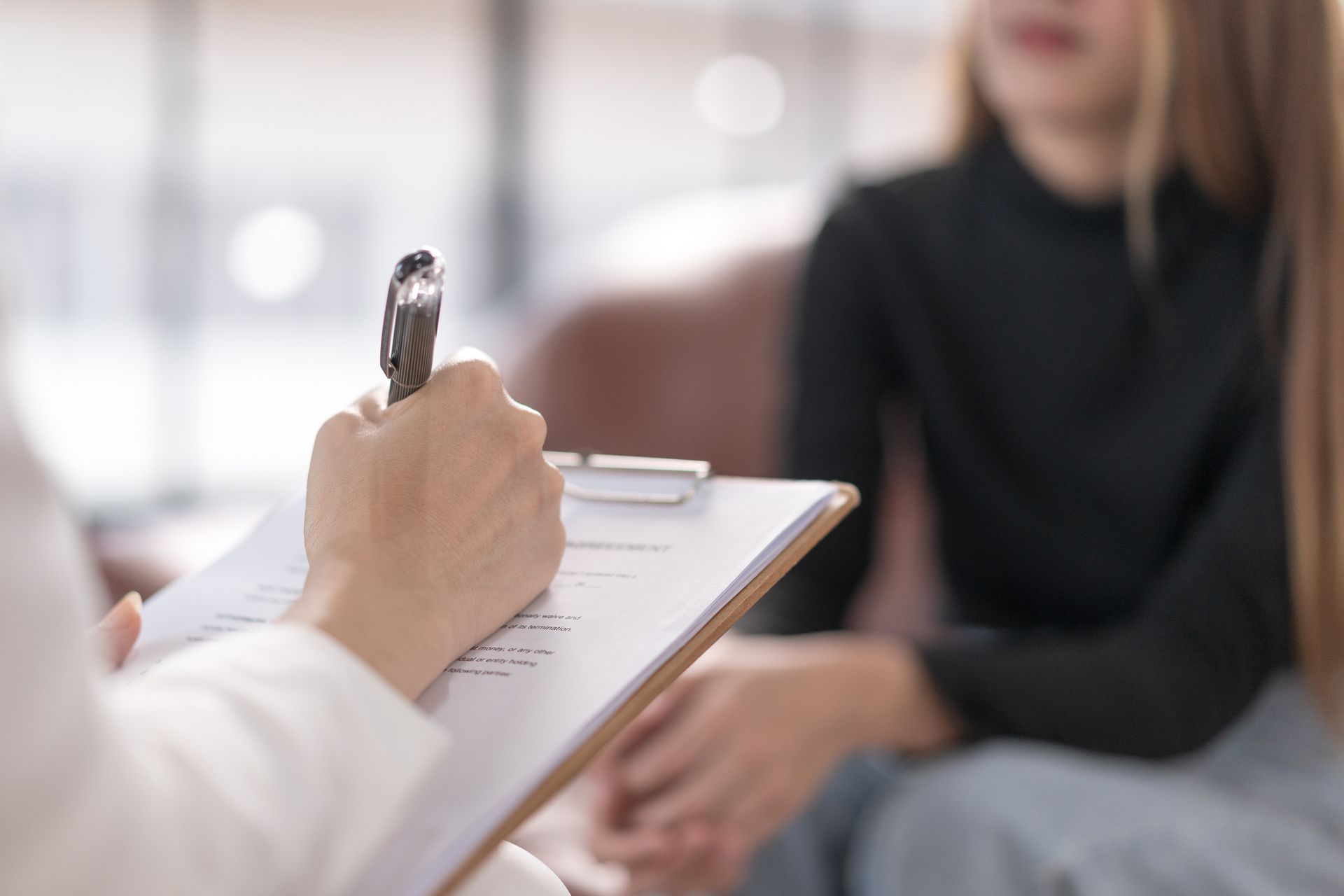 Clipboard with pen in foreground as a therapist talks with a seated client in a bright office