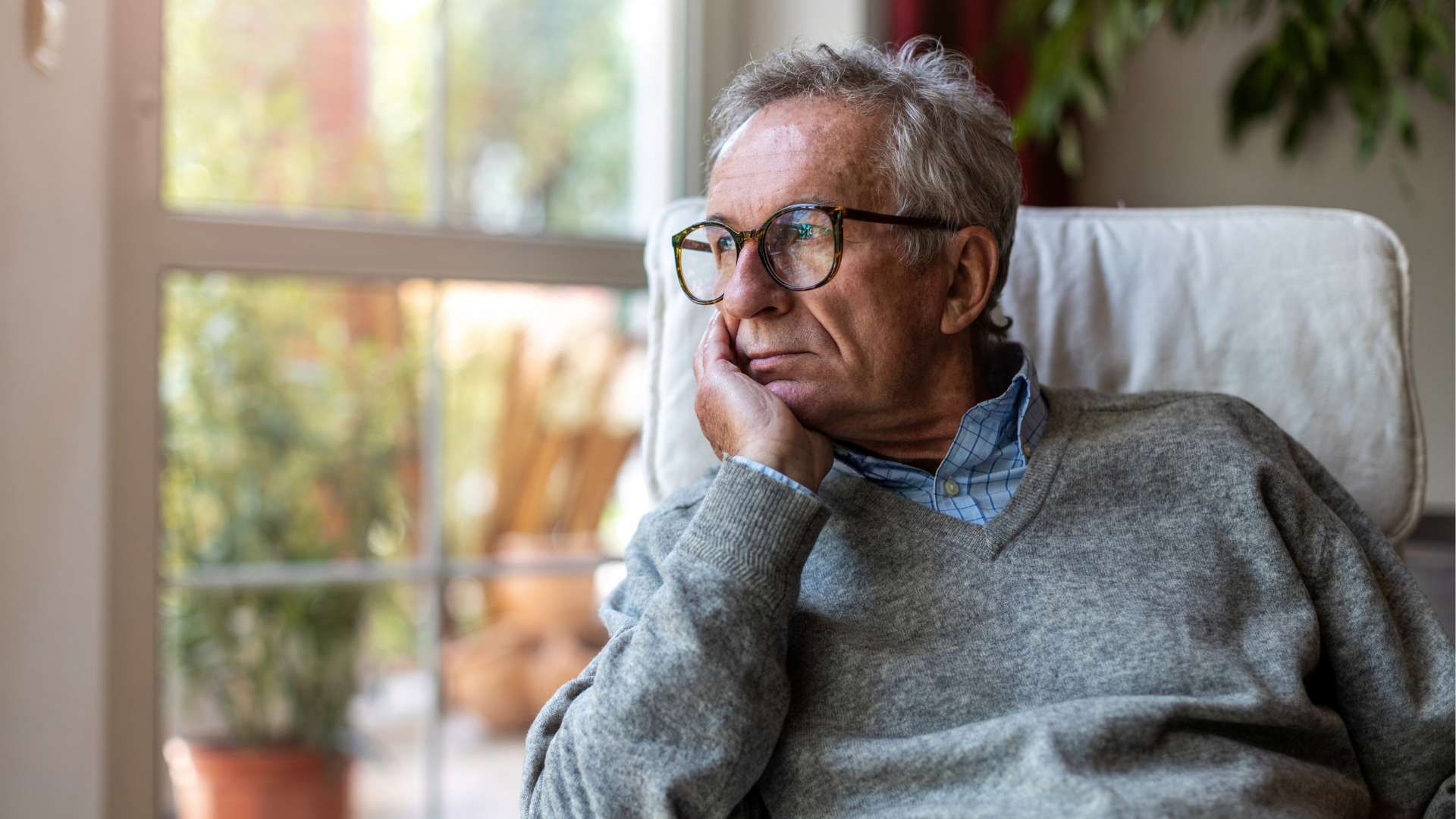 Older man in glasses sits by a window, resting his chin on his hand in a cozy living room.