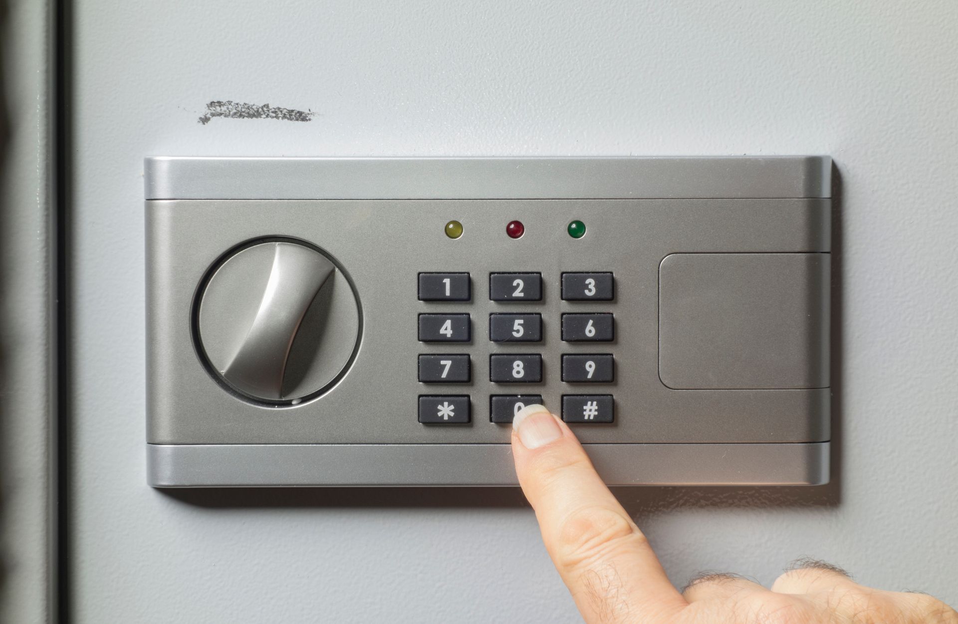 Hand pressing button 0 on a safe keypad. Safe is gray, with a combination dial and status lights.