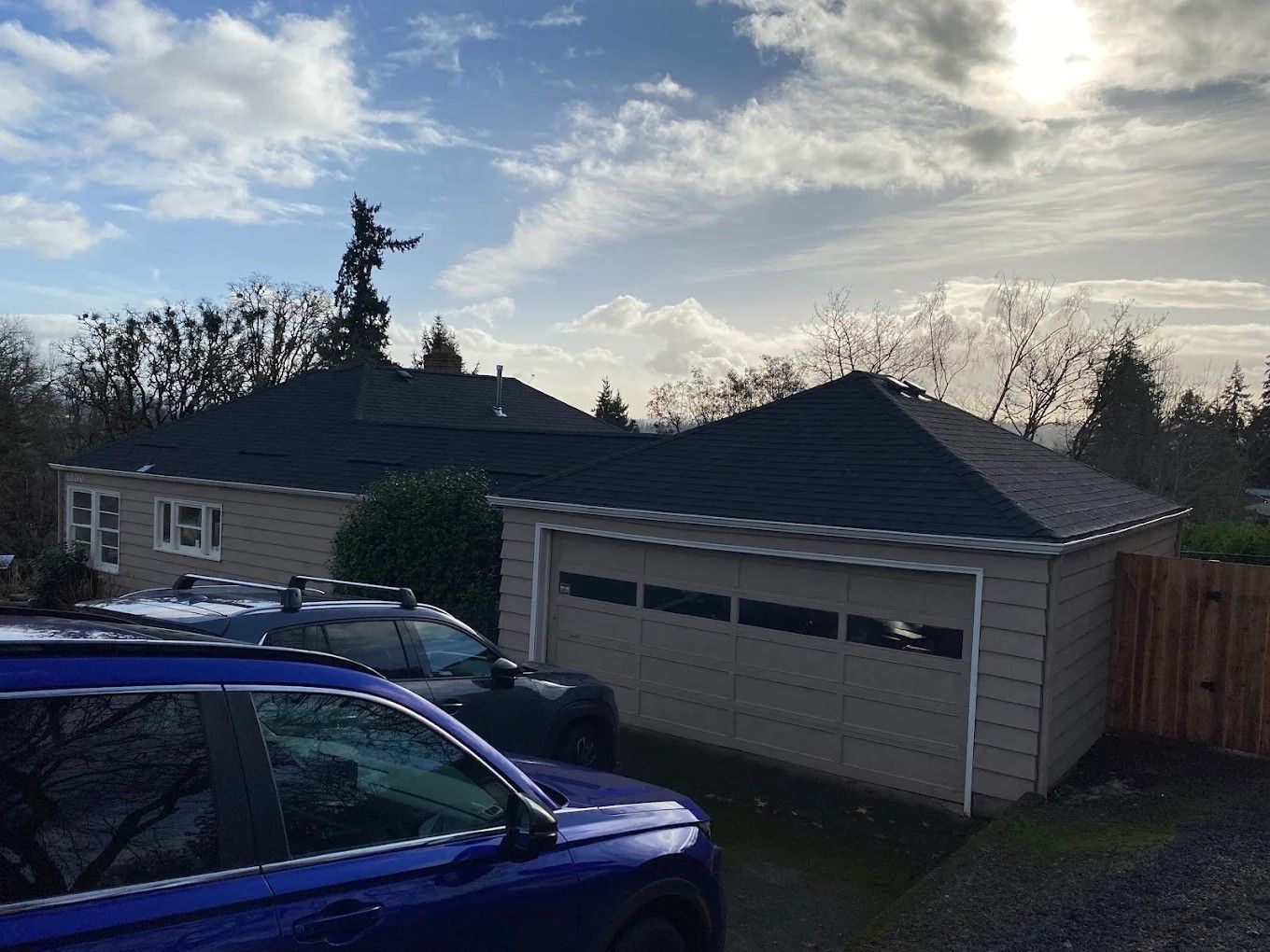 A tan house with a dark roof and a two-car garage, with a blue car parked in the driveway under a partly cloudy sky.