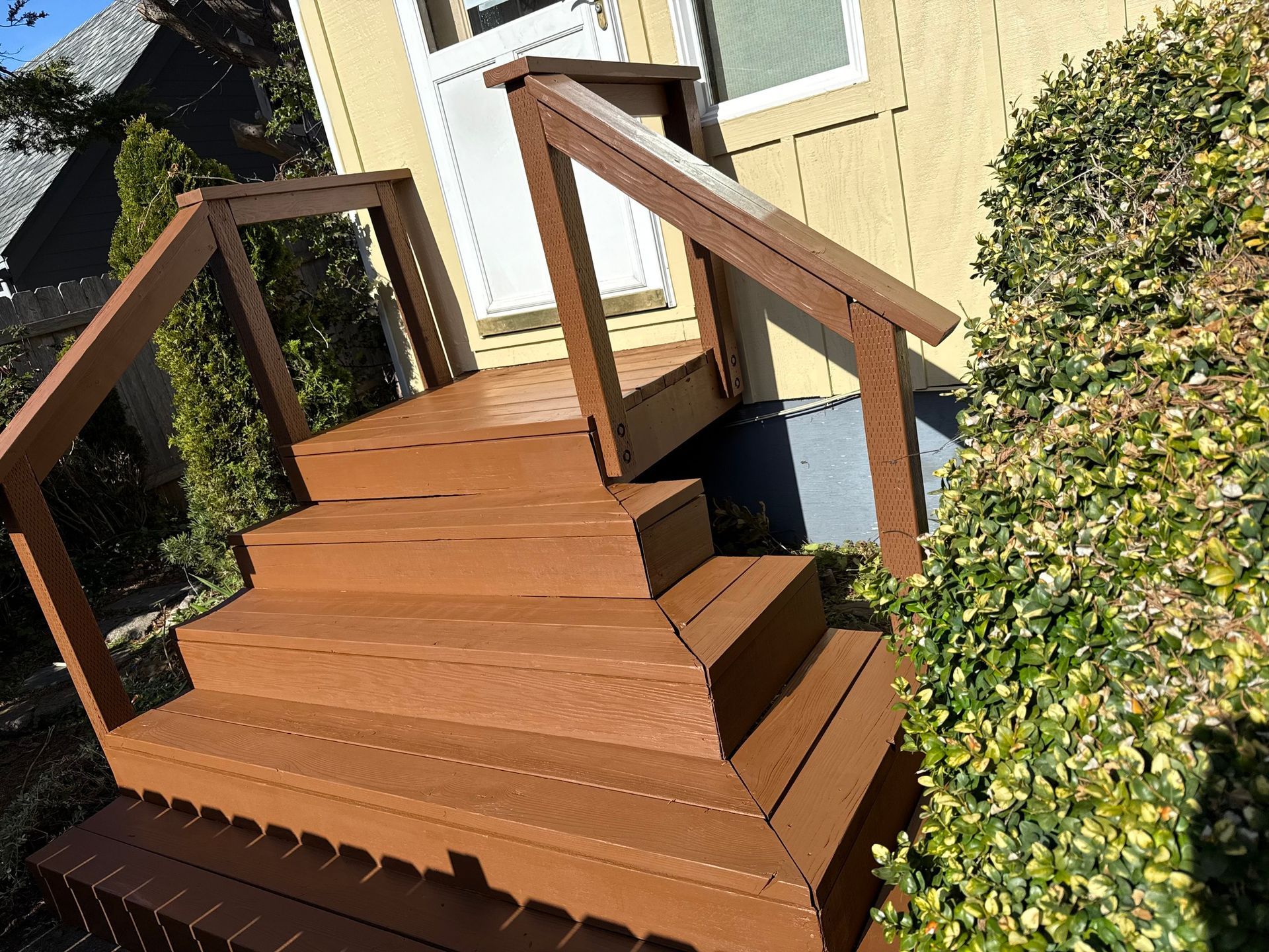 Wooden stairs with matching railings leading to an exterior doorway, situated next to a large shrub.