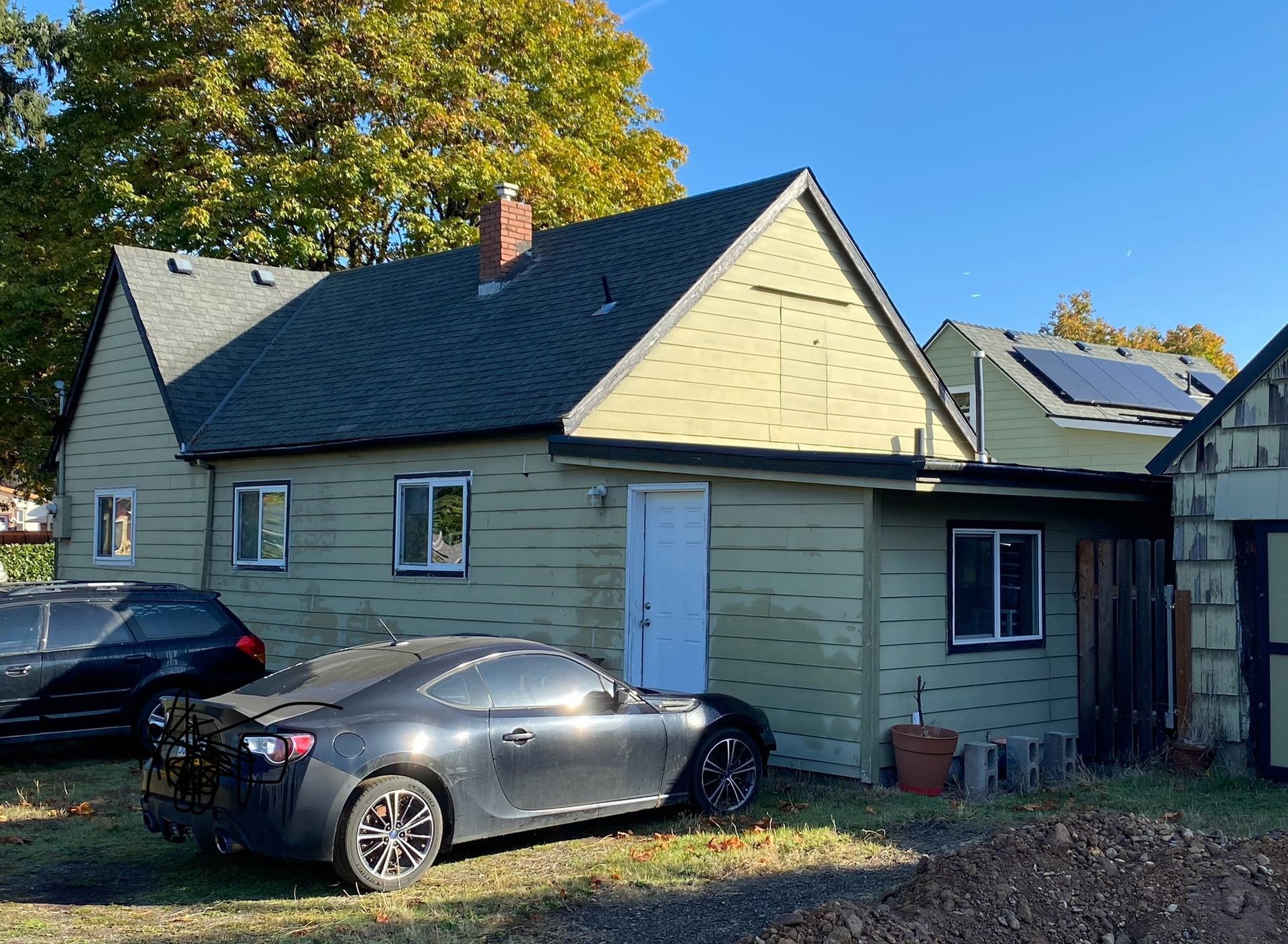 A pale green house with a dark roof and chimney, featuring a white door and two cars parked in the side yard.