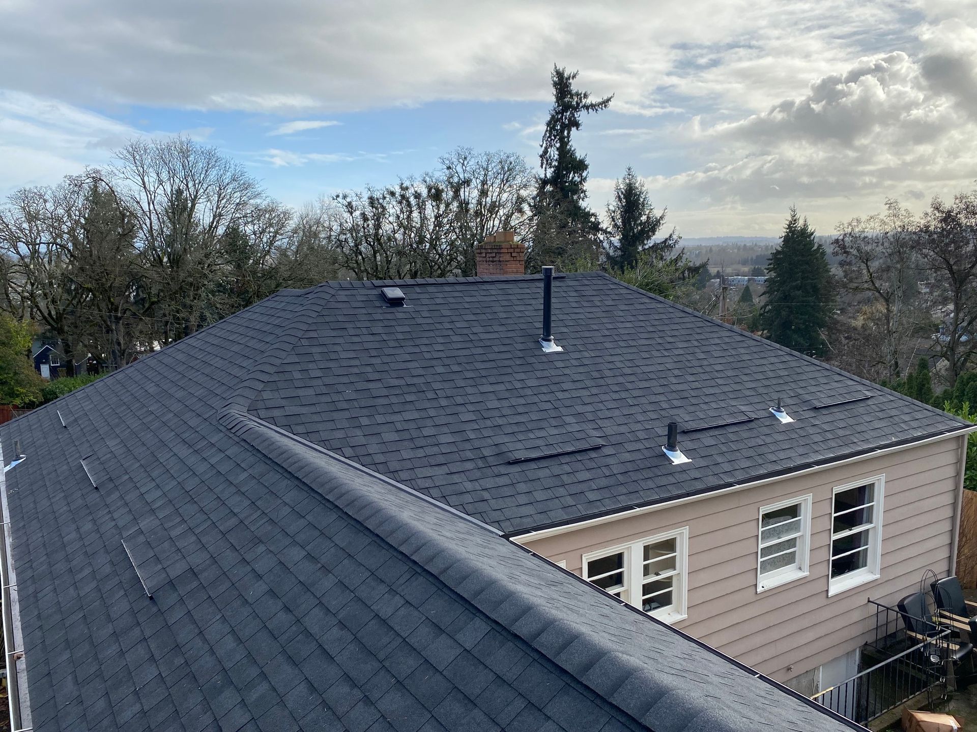 A high-angle view of a newly installed dark grey asphalt shingle roof on a house with light tan siding under a cloudy sky.