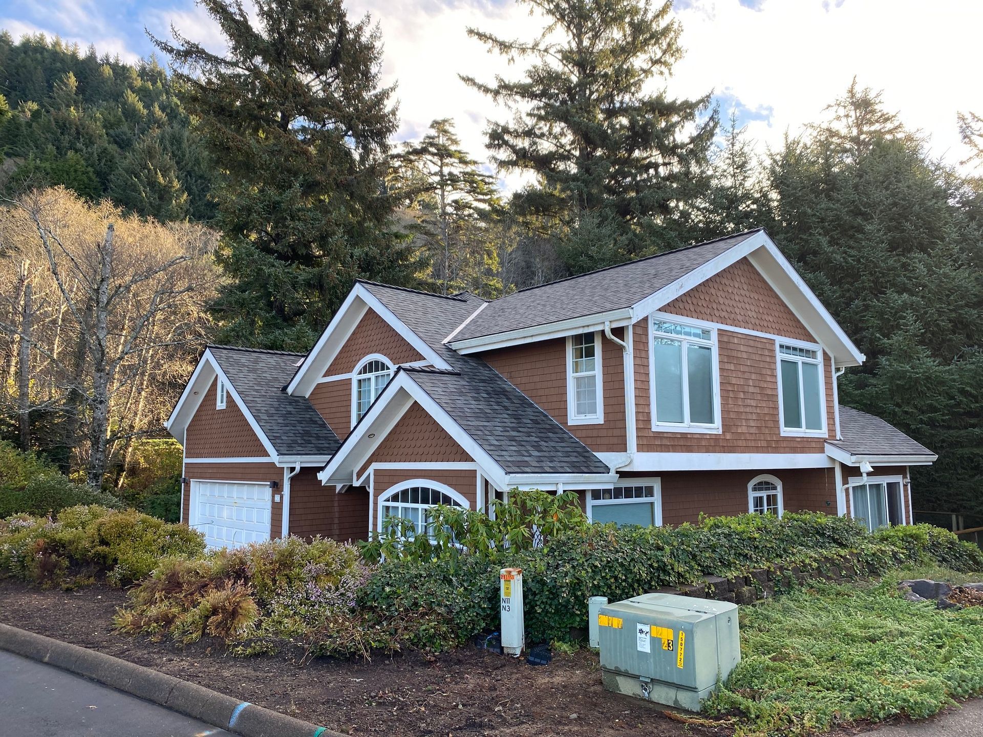 A two-story brown brick house with a gabled roof surrounded by trees, bushes, and an electrical box in the foreground.
