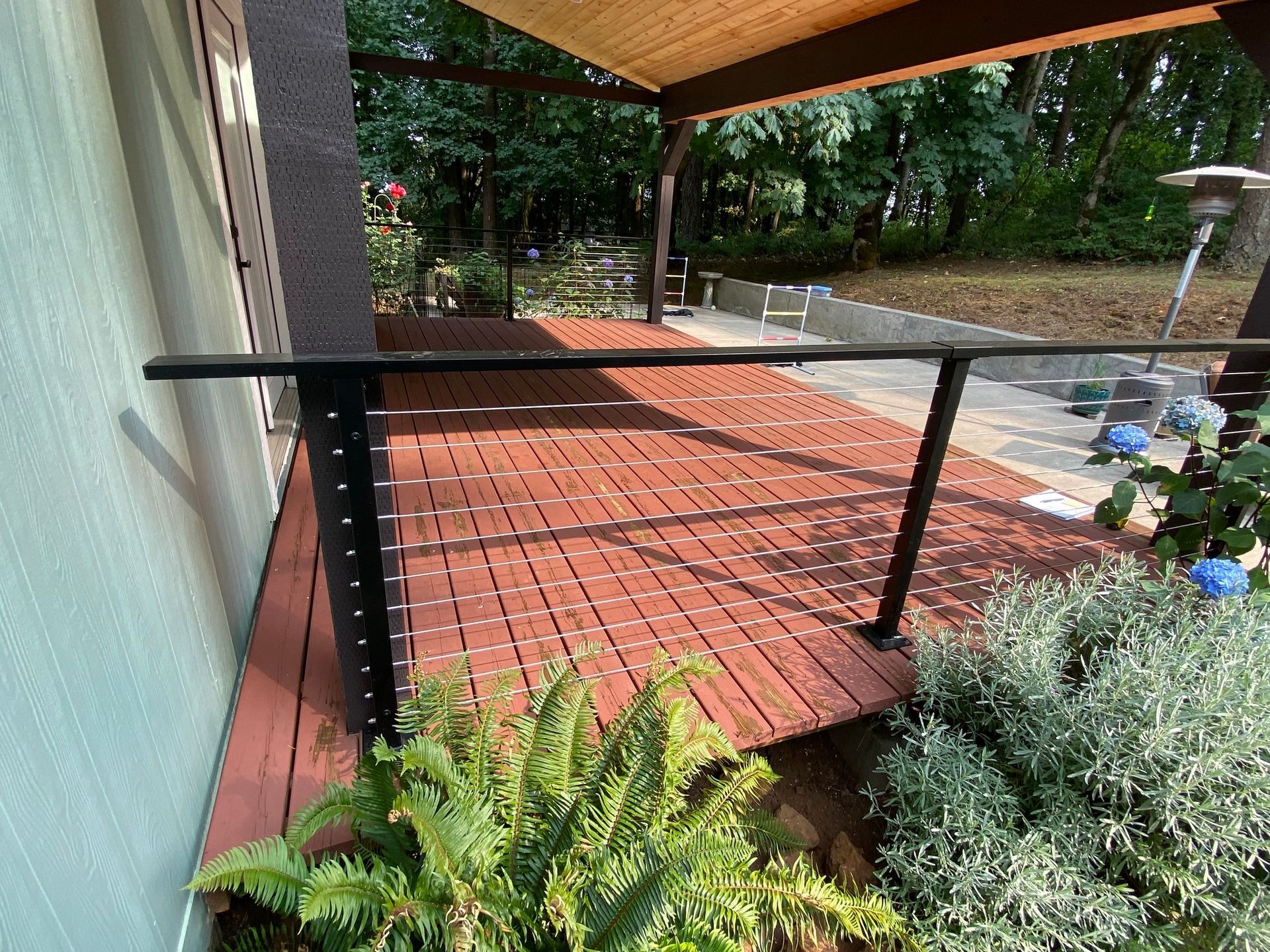 A red wood deck with a black metal cable railing, viewed from a patio level near lush green plants and trees.