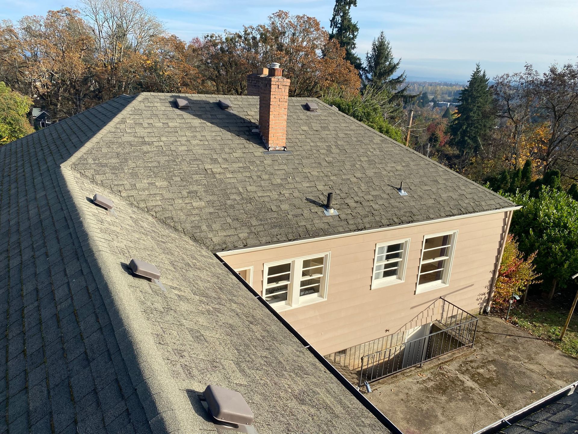 High-angle view of a beige house with a grey shingled roof, a brick chimney, and surrounding autumn trees.