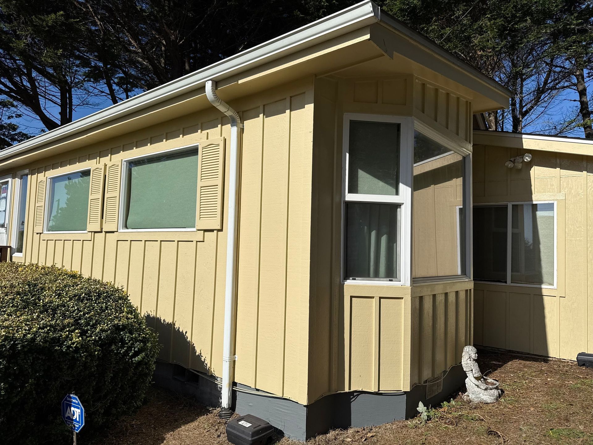 A yellow mobile home with vertical siding, bay windows, and a white downspout, set against a backdrop of trees.