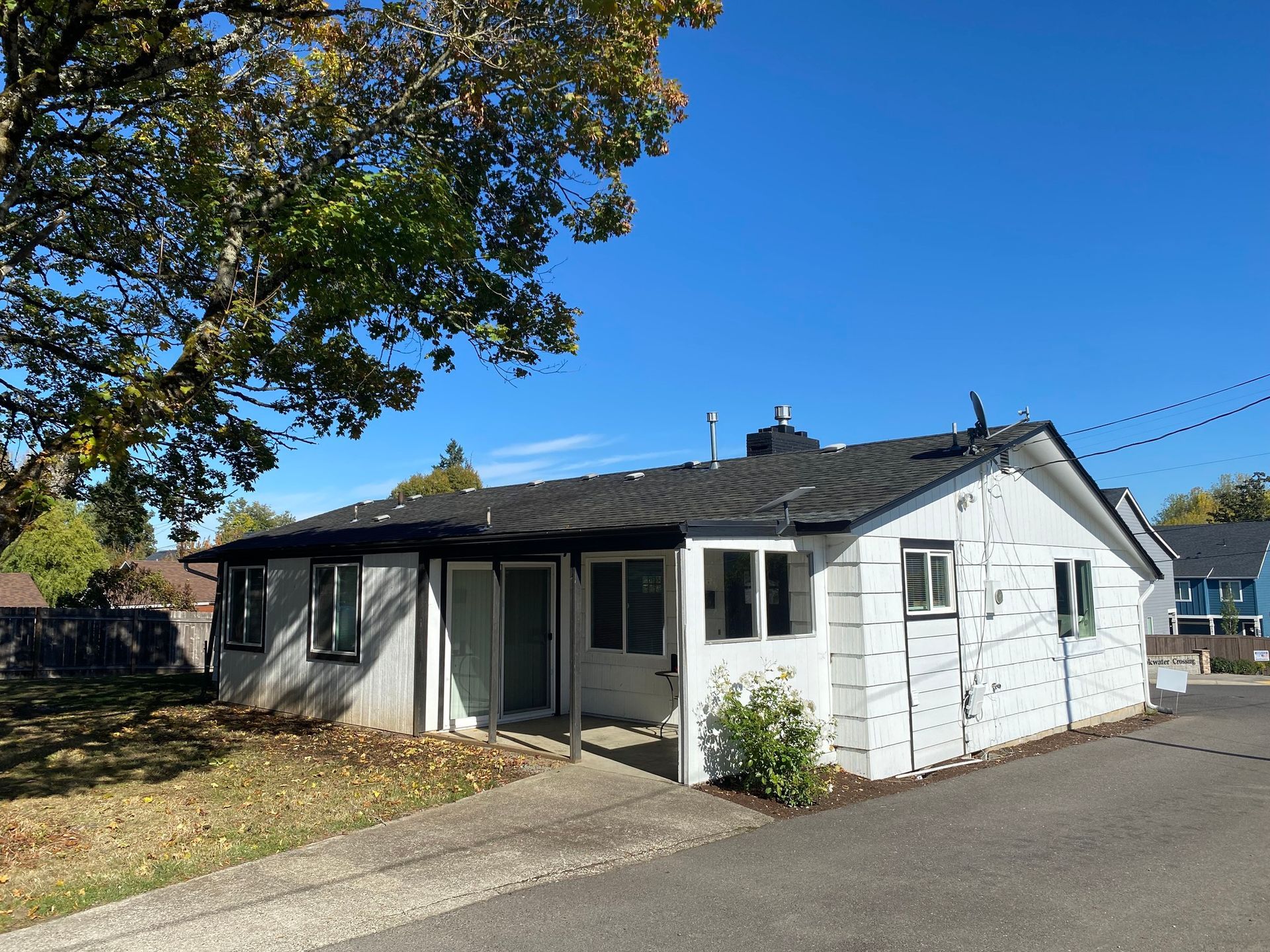 A white, single-story house with a black roof and a paved driveway under a clear blue sky.