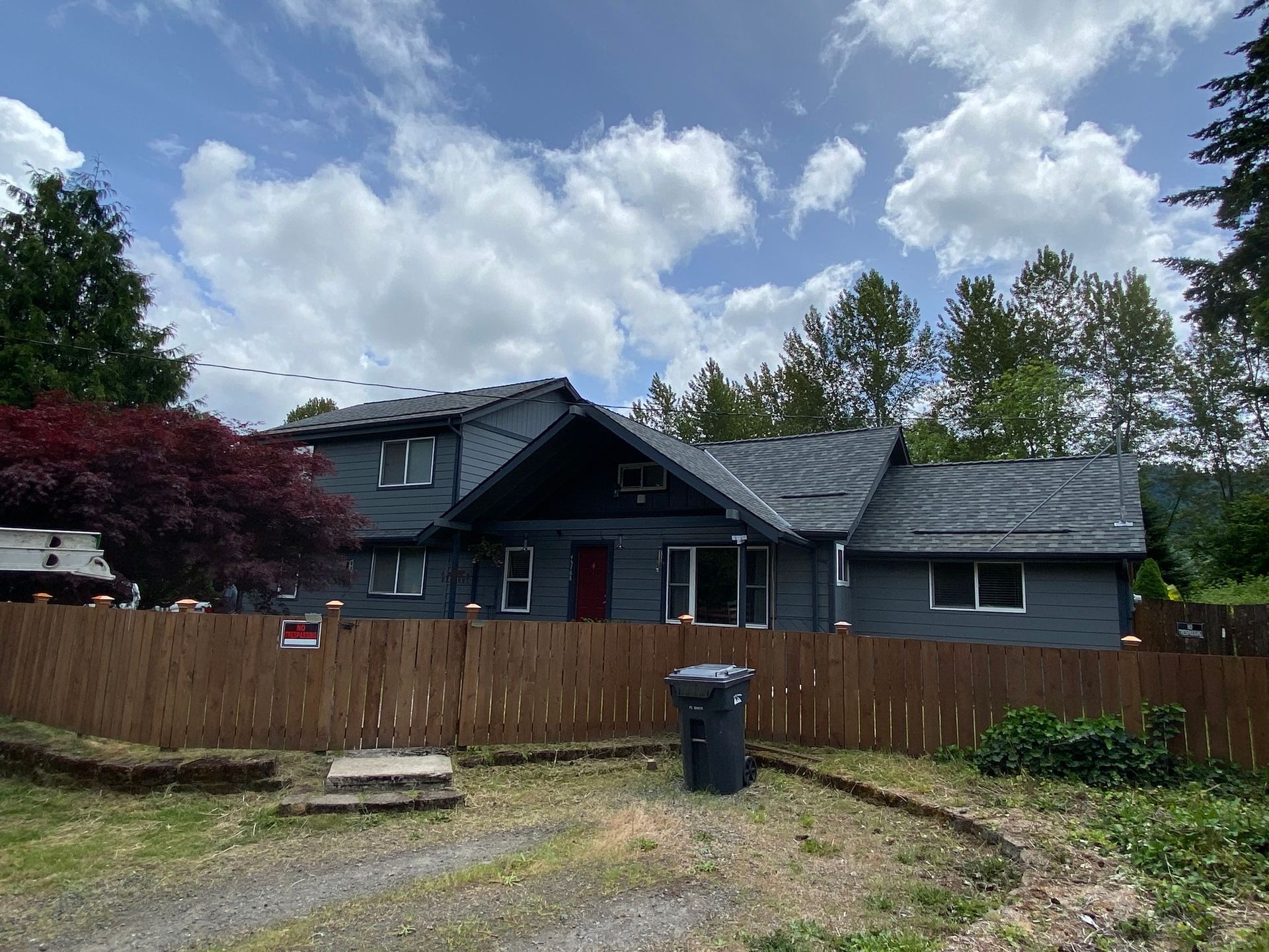 A two-story, dark gray house with a peaked roof and red front door, seen behind a tall wooden fence under a cloudy sky.