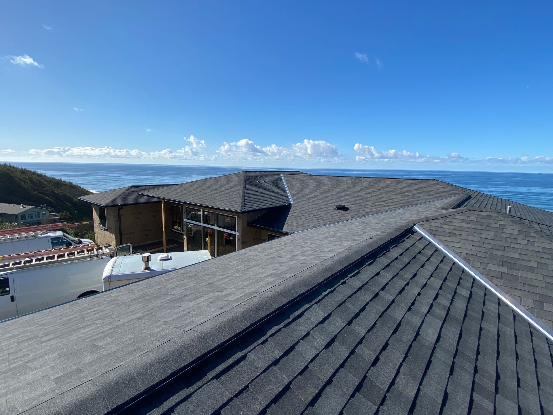A high-angle view of a grey shingled roof on a coastal home, overlooking the ocean under a clear blue sky.