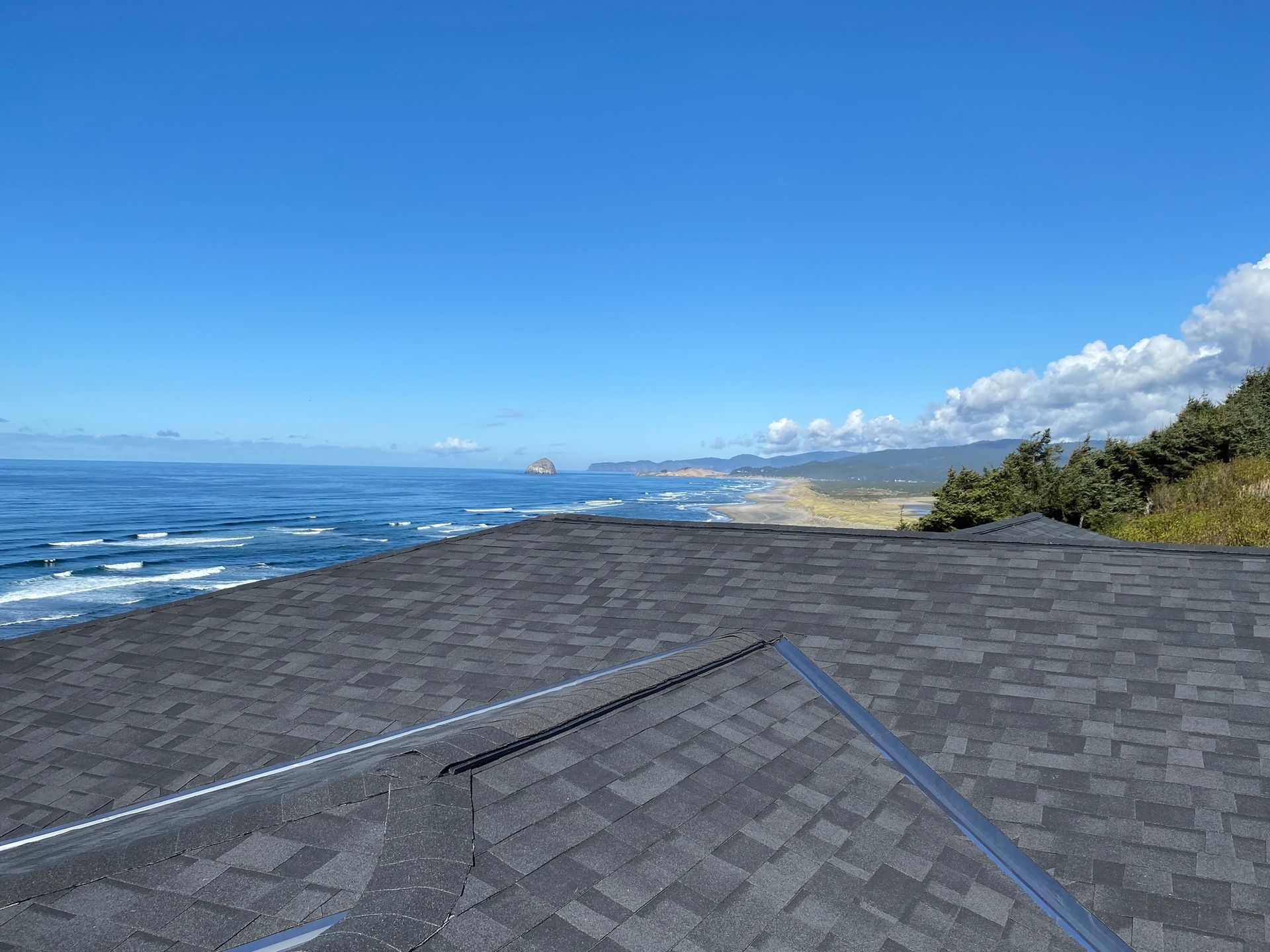 A high-angle view of a dark shingled roof overlooking a rocky coastline, ocean waves, and a sunny blue sky.