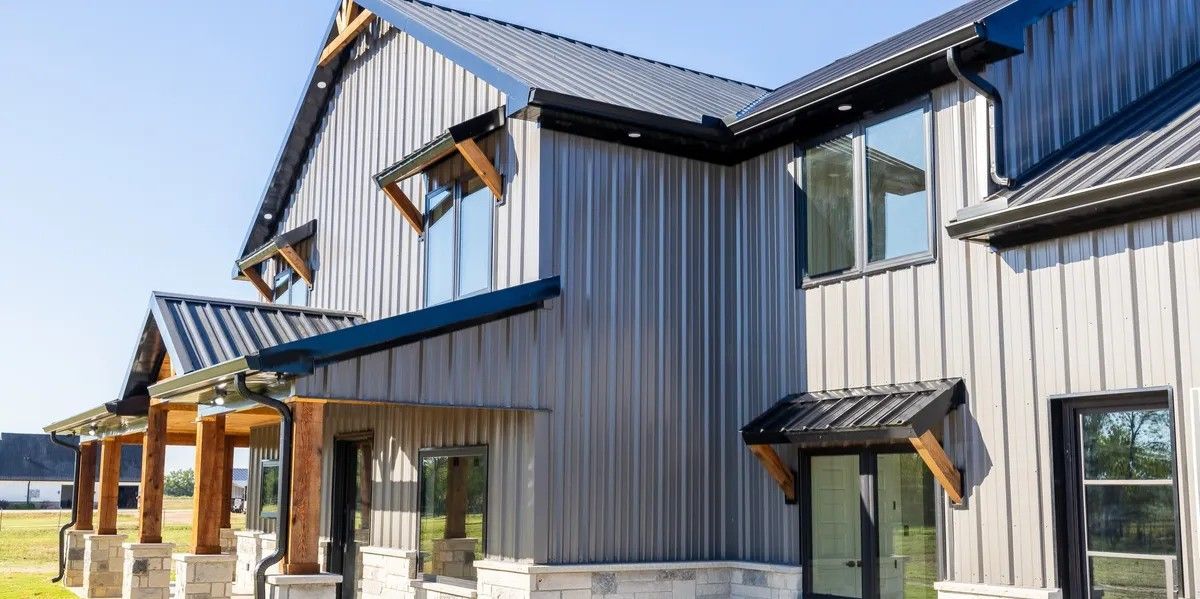 Modern house exterior with vertical gray metal siding, black roof, stone base, and wooden support beams on the porch.