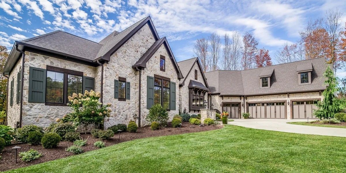 A large, two-story stone house with a multi-car garage, dark shutters, and a lawn under a blue, cloudy sky.