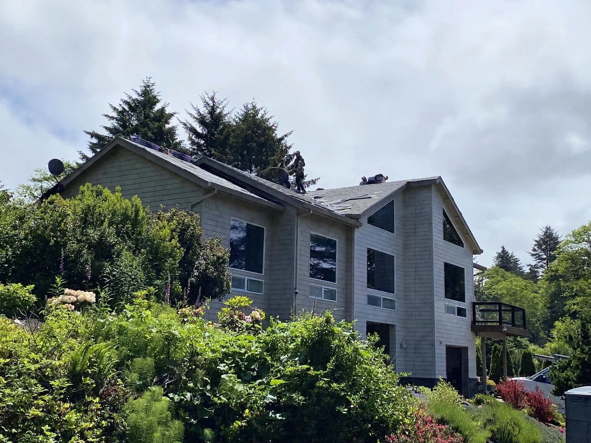 A two-story, light-colored house with a dark roof nestled among green trees and bushes under a cloudy sky.