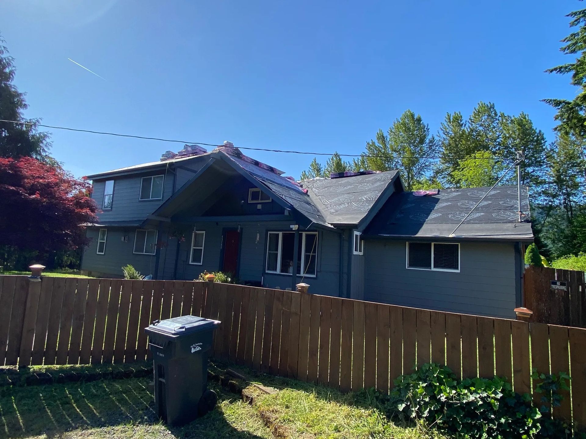 A two-story grey house with a roof under repair, behind a wooden fence and a trash bin under a clear blue sky.