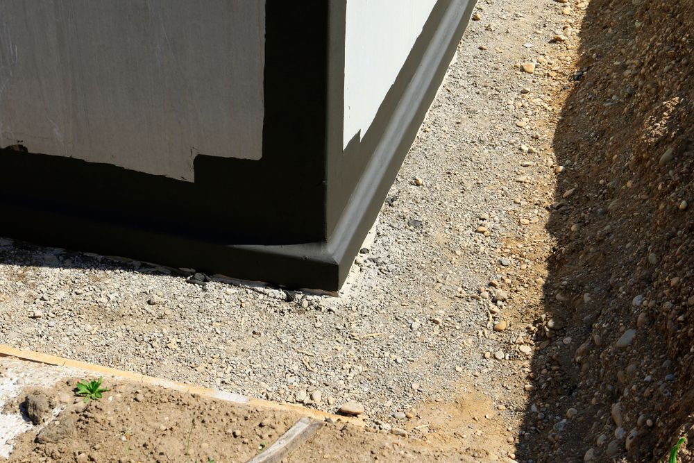 A building corner with a black-coated foundation, surrounded by a gravel drainage strip next to an unpaved earthen bank.
